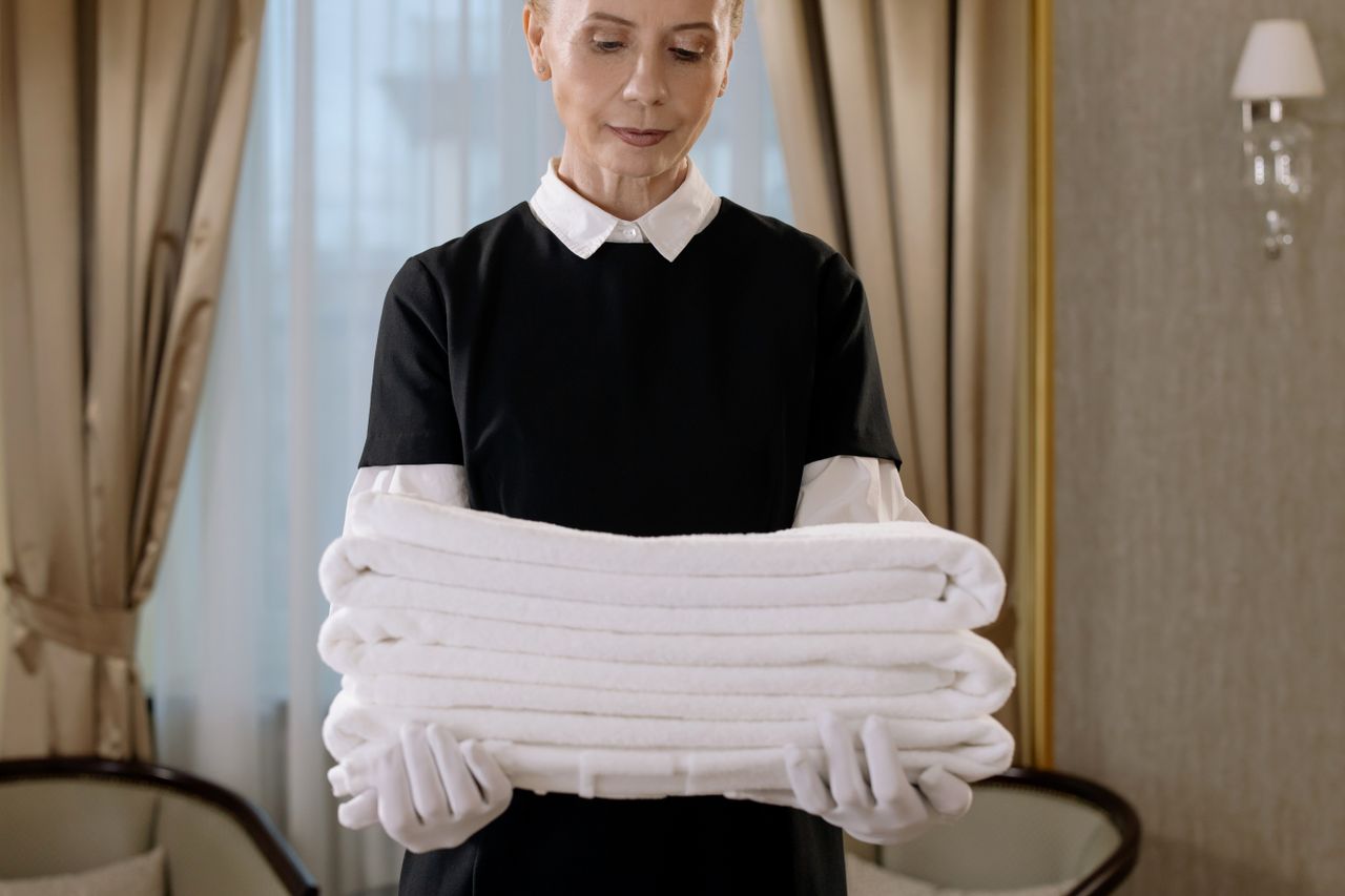 hero-02 Professional housekeeper holding a stack of fresh towels in a luxurious hotel room, showcasing elegance and service.