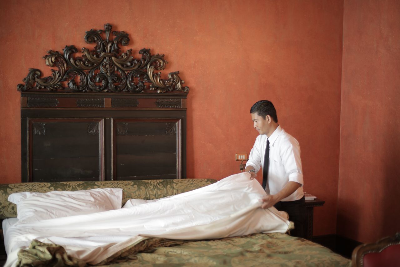 hero-01 A hotel staff member in formal attire making a bed in a luxurious bedroom setting.