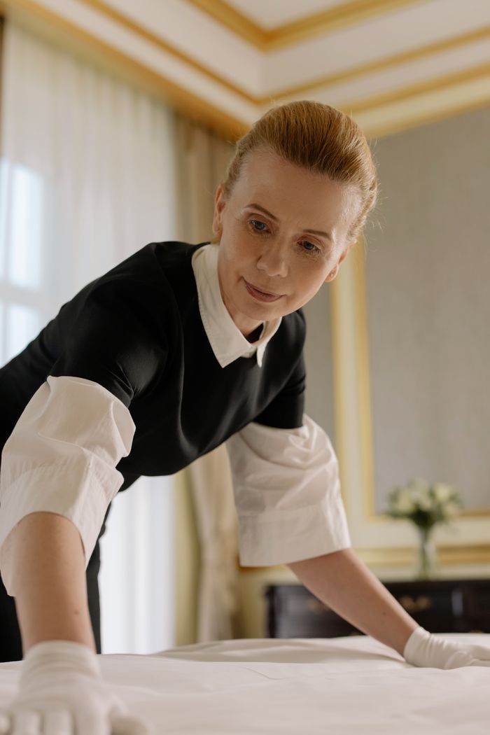 services-02 A housekeeper in uniform skillfully arranges bed linen in a luxurious hotel room.